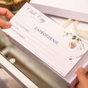 Dłonie trzymające eleganckie zaproszenie upominkowe z delikatną typografią i kwiatowymi wzorami.